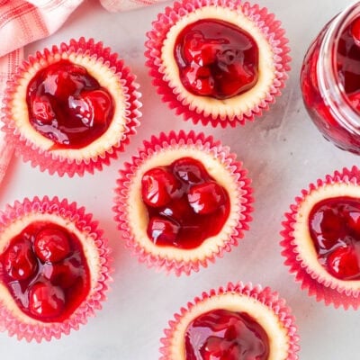 An overhead shot of six mini cherry cheesecakes next to a jar of cherries.