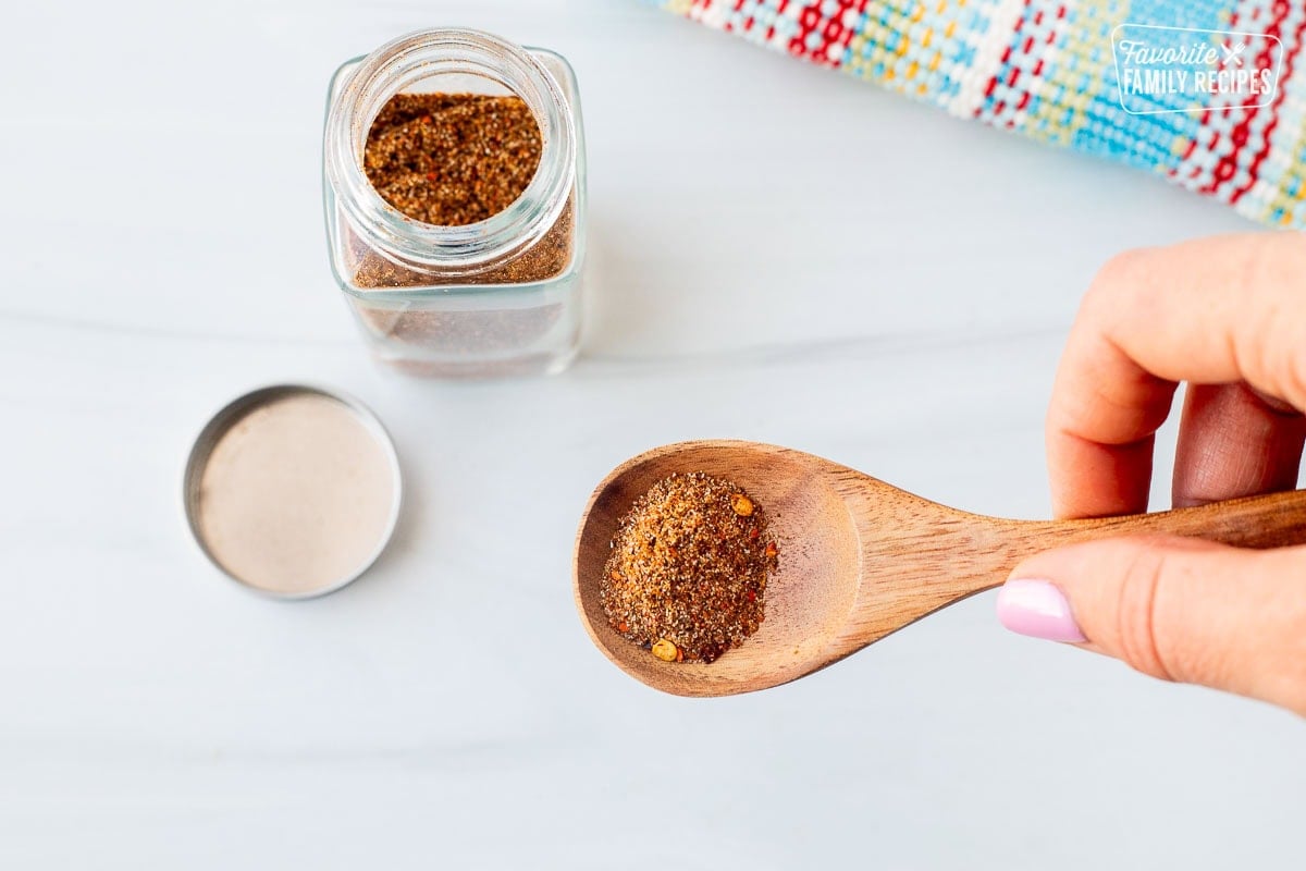 Hand holding up a spoon fill of Carne Asada seasoning. Small glass jar of Carne Asada seasoning on the side.
