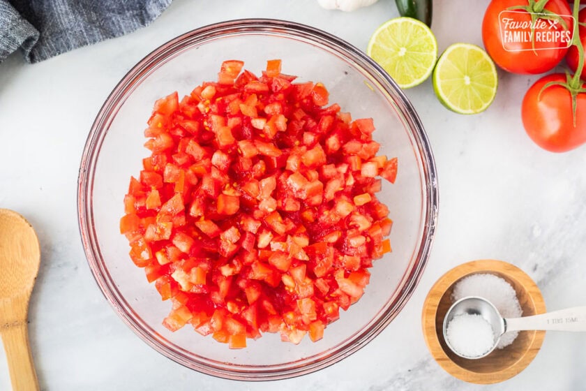 Glass mixing bowl with diced tomatoes next to bowl of kosher salt.