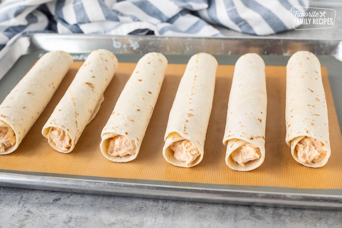 Six rolled chicken flautas on a baking sheet.