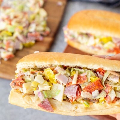 Holding a chopped Italian sandwich with lettuce, pepperoni, salami, sliced ham, provolone cheese, diced red onion, mayo, pickles and banana peppers.