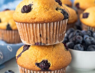 Two blueberry muffins stacked on top of each other next to cooling rack of blueberry muffins.
