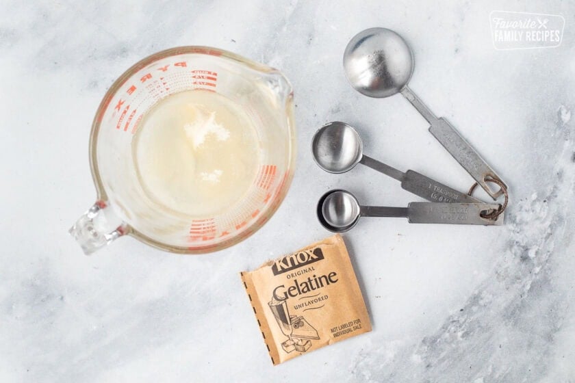 Unflavored gelatin in a glass dish combined with water and measuring spoons.