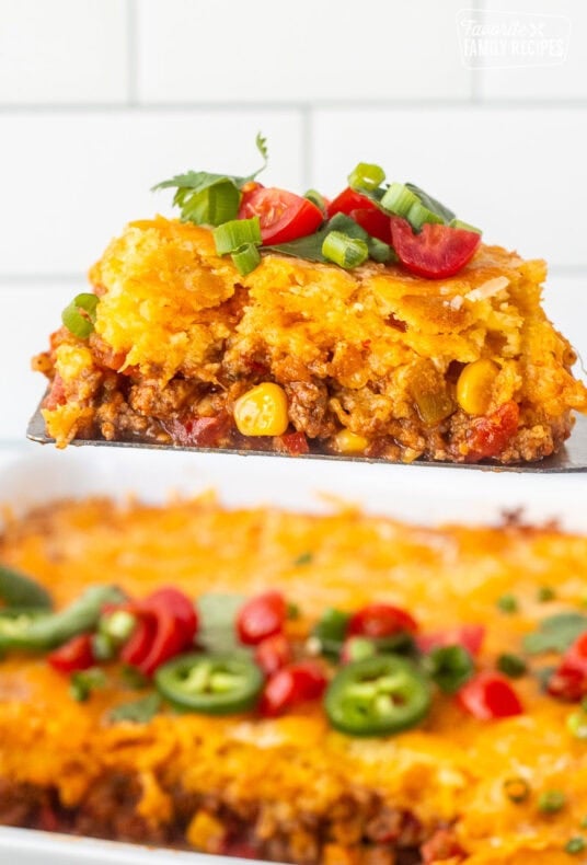 Spatula holding a slice of Mexican cornbread casserole topped with fresh cilantro, diced tomatoes and jalapeños.