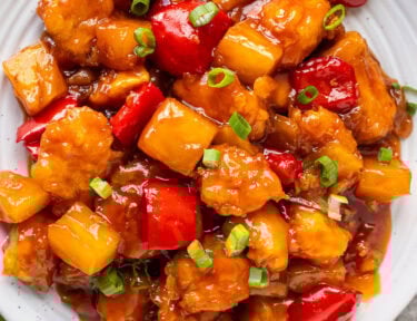 Sweet and sour chicken garnish with sliced green onions on a plate. Bowl of rice on the side.