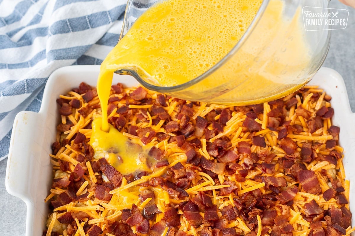 Pouring egg mixture on top of bacon, cheddar cheese and tater tots in a baking dish.