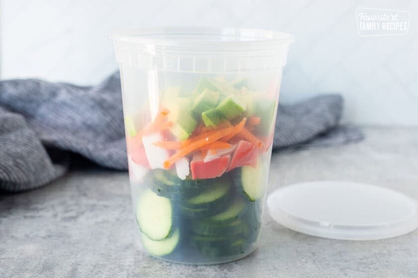 Plastic container with sliced cucumbers, cut imitation crab, carrot and avocado.
