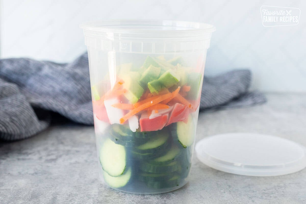 Plastic container with sliced cucumbers, cut imitation crab, carrot and avocado.