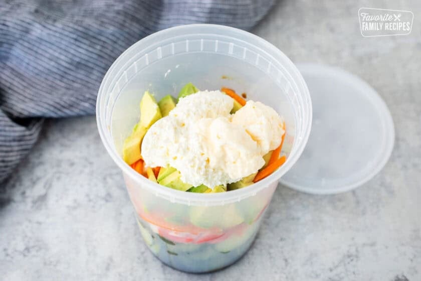 Plastic container with sliced cucumbers, cut imitation crab, carrot and avocado. Mayo and whipped cream cheese on top.
