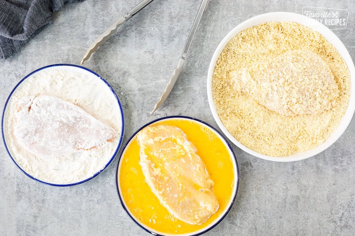 Chicken breasts in a plate of flour, plate of egg wash and bowl of bread crumbs.