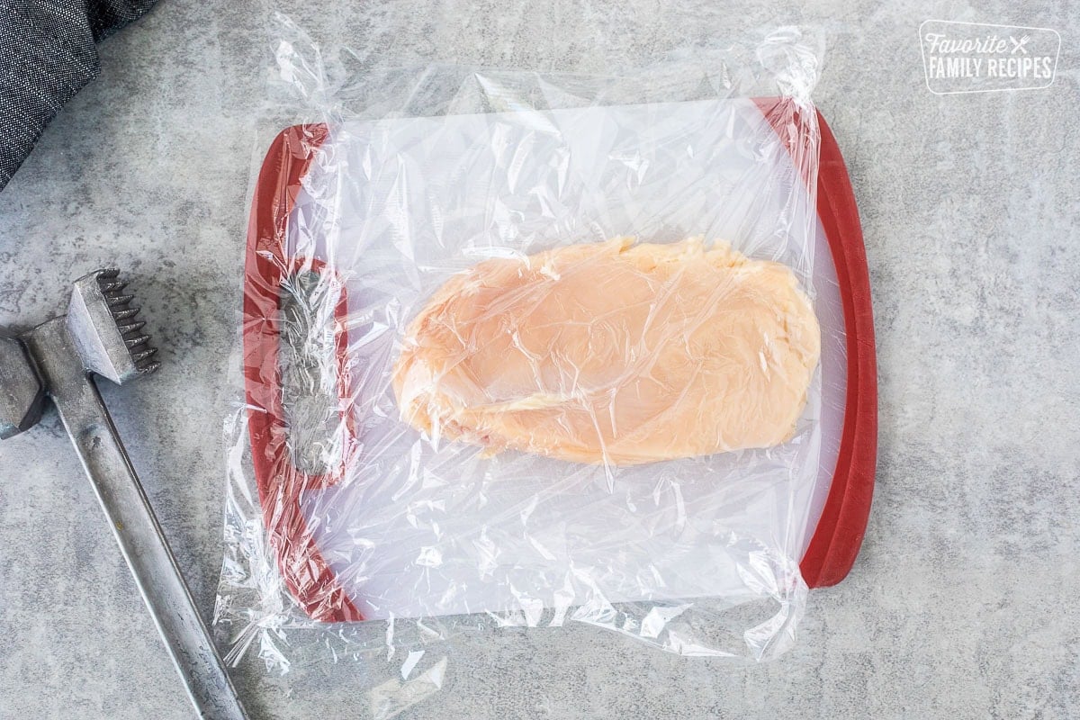 Cutting board with flattened chicken breast covered in plastic wrap and a metal mallet on the side.