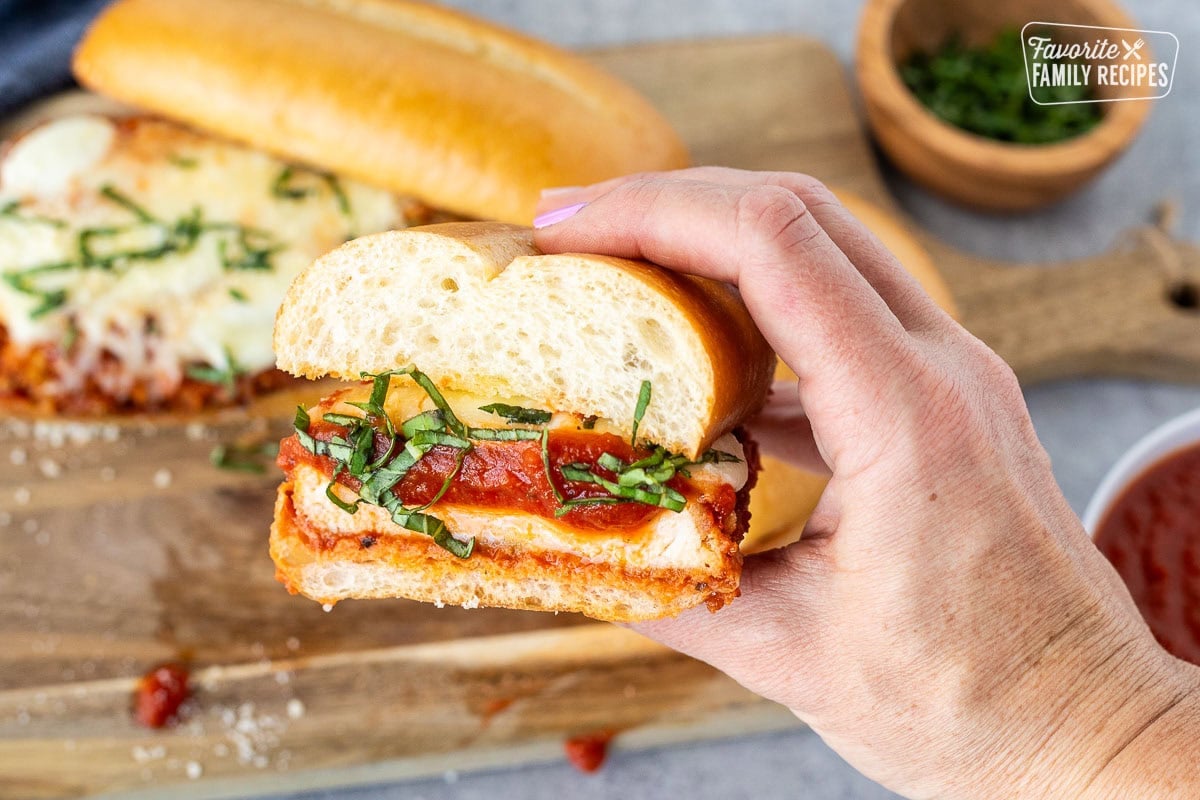 hand holding a Chicken Parmesan Sandwich cut in half with marinara, melted mozzarella cheeses and fresh basil for garnish.