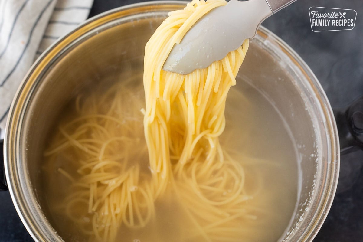 Cooked spaghetti pasta held up with tongs out of a large pot of hot water.