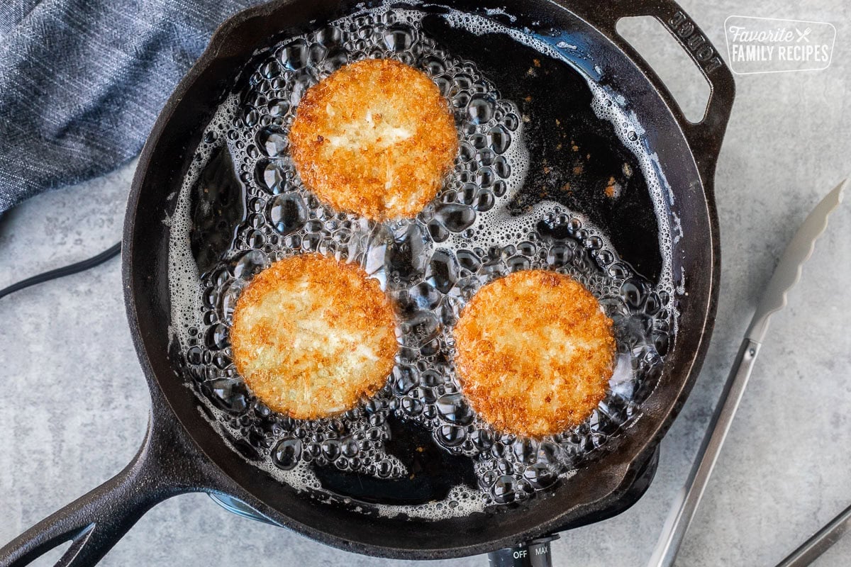 Three crab cakes frying in a cast iron skillet with hot oil. Tongs on the side.