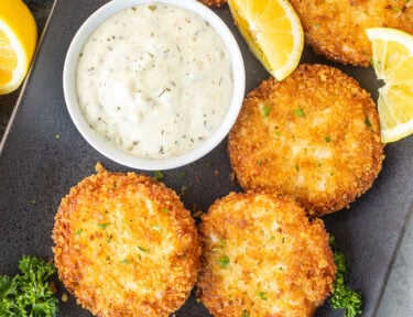 Golden crab cakes on a platter with a small bowl of tartar sauce.
