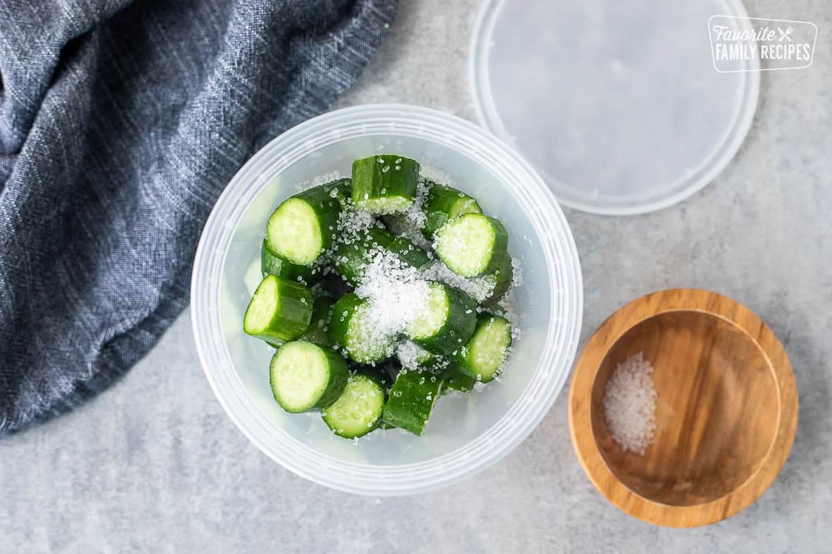 Plastic container with chopped baby cucumbers and salt. Plastic lid on the side. Wooden bowl of salt.