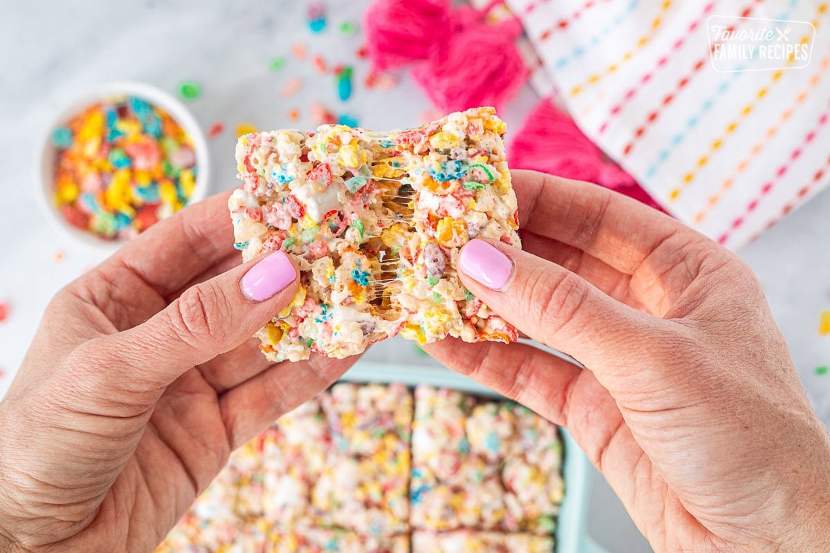 Hands stretching apart a gooey marshmallow fruity pebbles rice crispy treat.