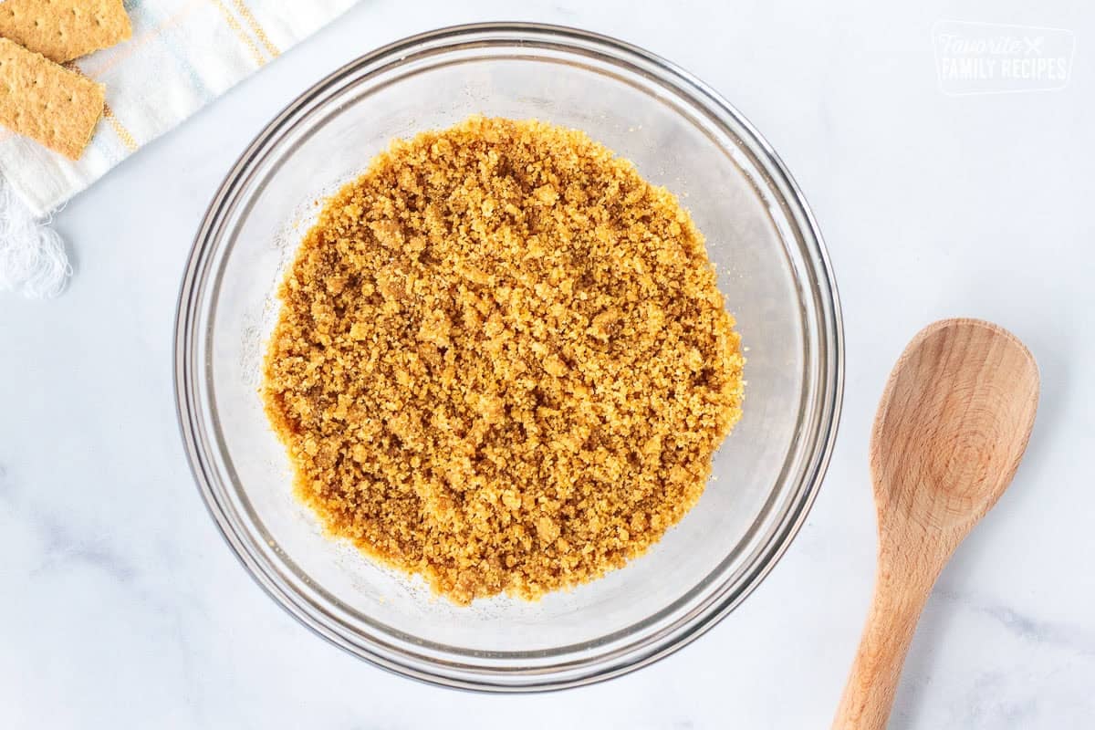 Glass bowl with crushed graham crackers combined with butter and sugar. Wooden spoon on the side.