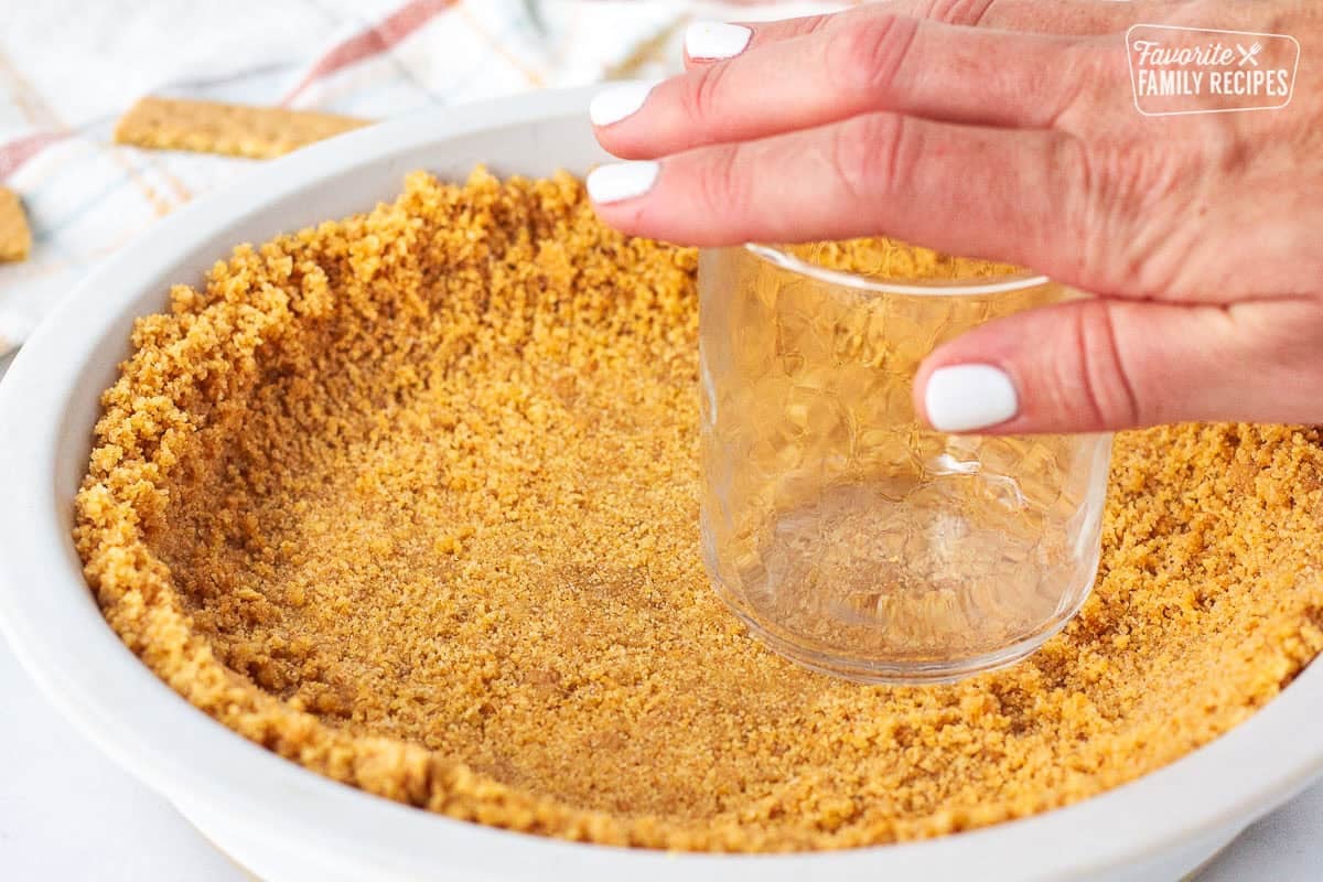 Using a glass to p down graham cracker pie crust by hand into a pie plate.