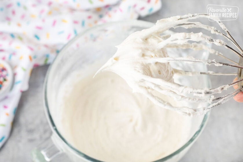 Whisk with frosting over a glass mixing bowl of frosting.