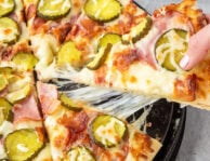 Pulling a piece of cheesy Pickle pizza with ranch, mozzarella cheese, Canadian bacon, bacon and pickle chips. Pickle chips on the side.