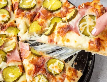 Pulling a piece of cheesy Pickle pizza with ranch, mozzarella cheese, Canadian bacon, bacon and pickle chips. Pickle chips on the side.