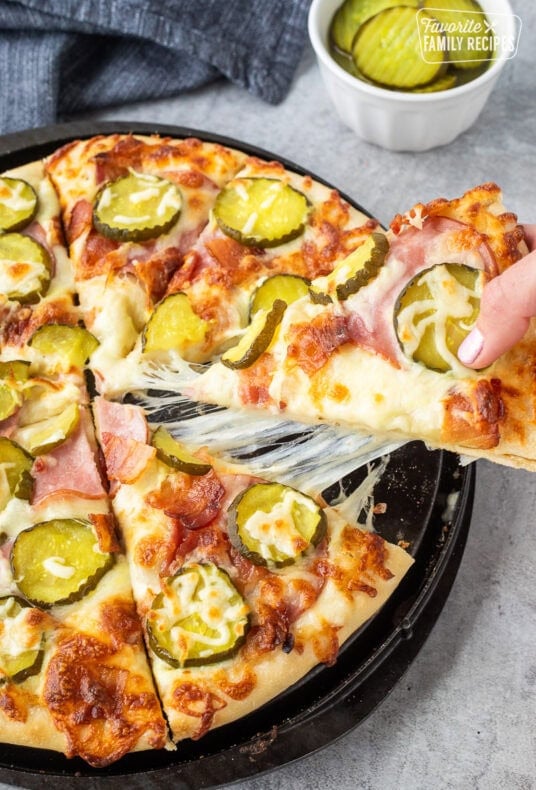 Pulling a piece of cheesy Pickle pizza with ranch, mozzarella cheese, Canadian bacon, bacon and pickle chips. Pickle chips on the side.