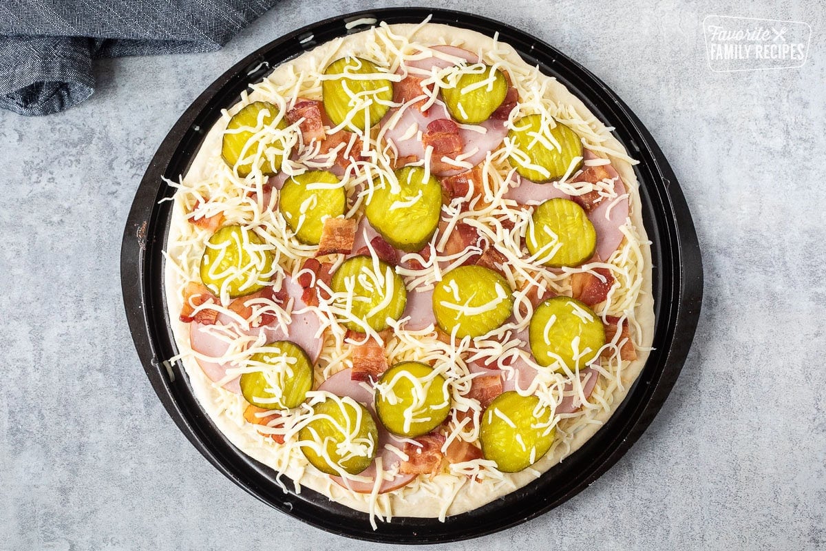 Unbaked Pickle pizza with ranch, mozzarella cheese, Canadian bacon, bacon and pickle chips with extra mozzarella cheese on top.