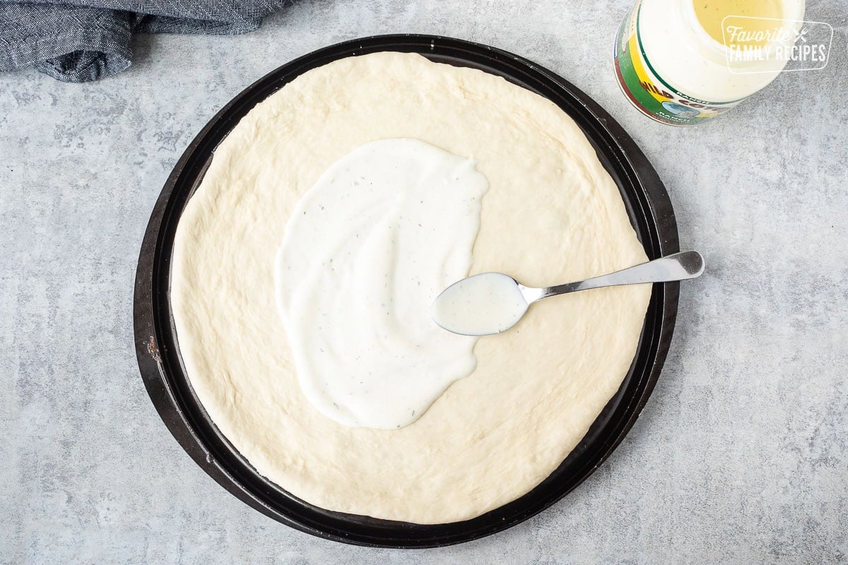 Spreading Ranch dressing with a spoon on a rolled out pizza dough.