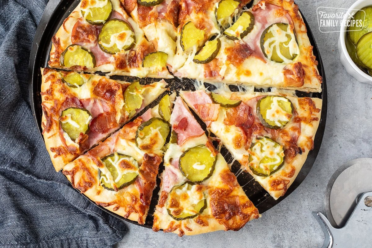 Cut Pickle pizza with ranch, mozzarella cheese, Canadian bacon, bacon and pickle chips. Pizza cutter and pickle chips on the side.