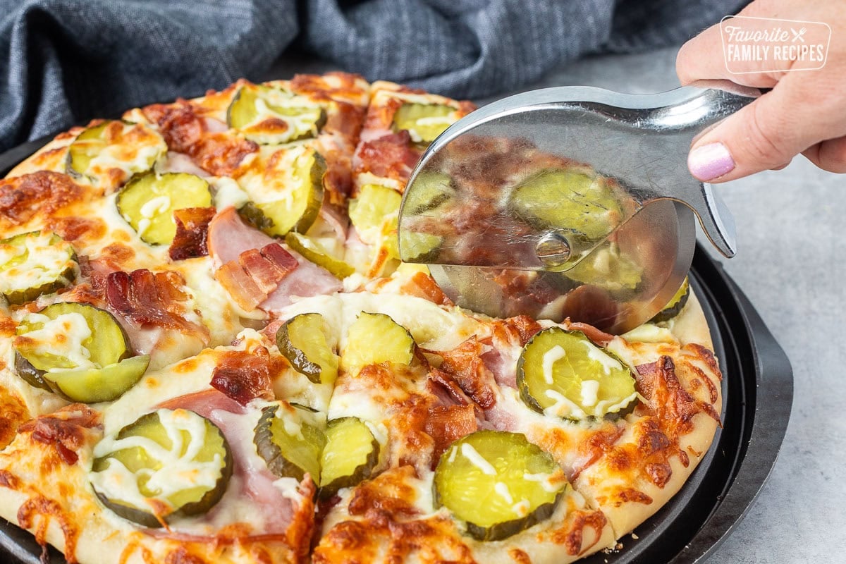 Cutting a Pickle Pizza with a pizza slicer with with ranch sauce, mozzarella cheese, Canadian bacon, bacon and pickle chips.