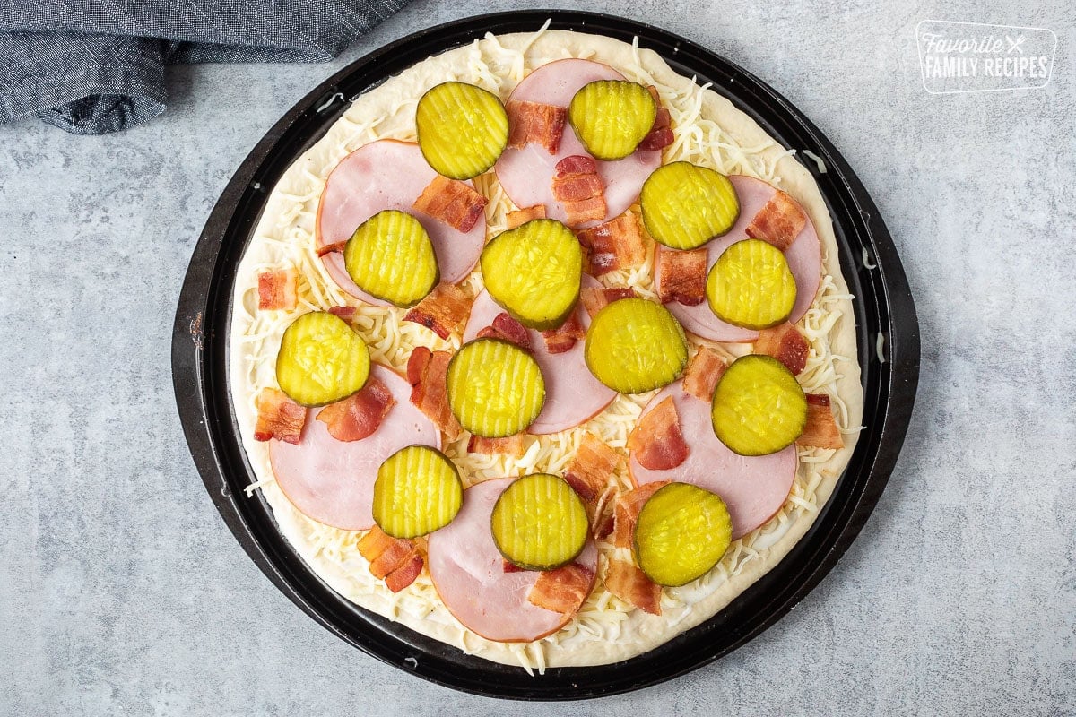 Unbaked Pickle pizza with ranch, mozzarella cheese, Canadian bacon, bacon and pickle chips.