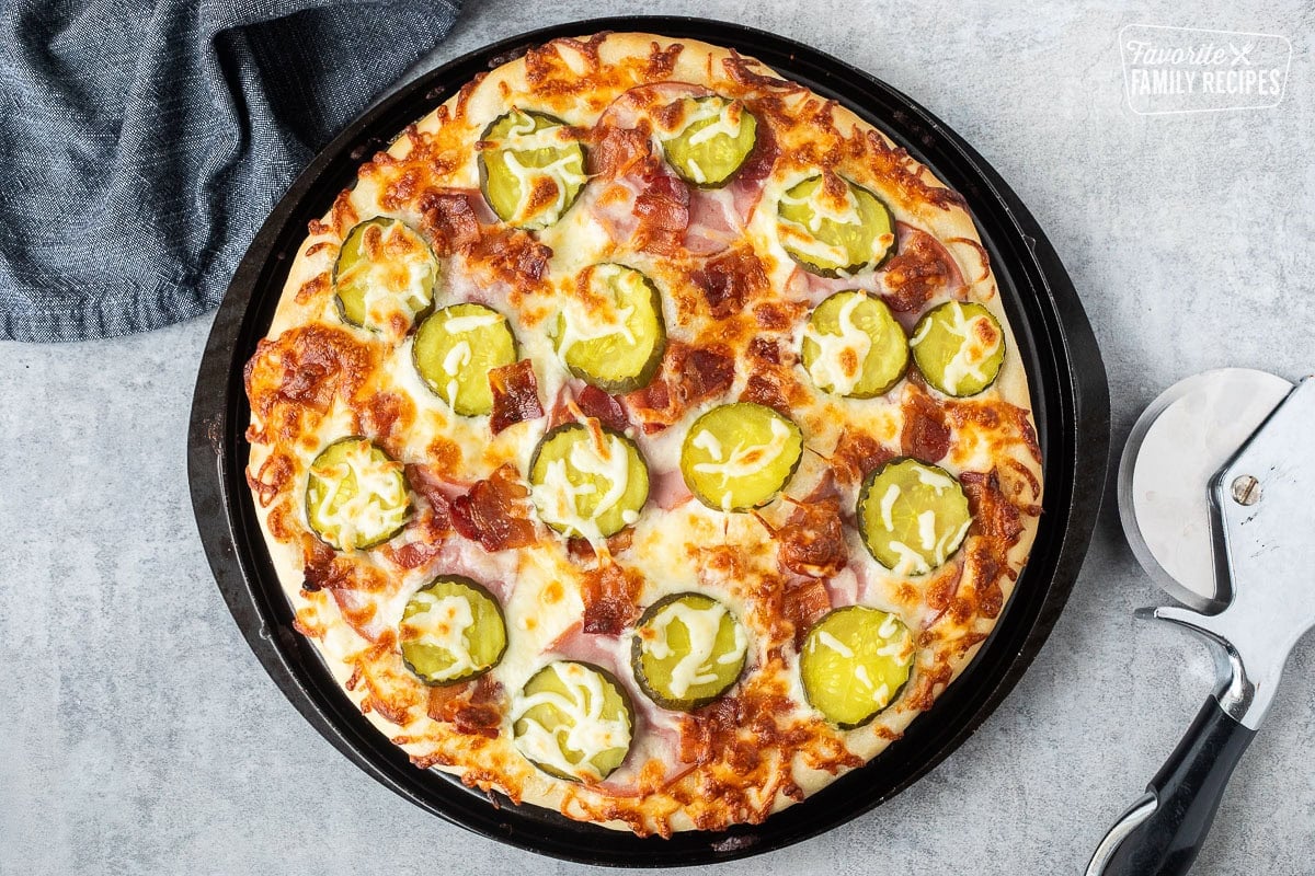 Baked Pickle pizza with ranch, mozzarella cheese, Canadian bacon, bacon and pickle chips. Pizza cutter on the side.