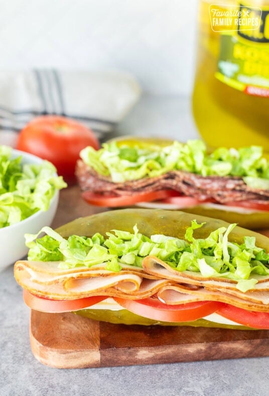 Two pickle sandwiches on a cutting board with turkey, lettuce, tomato, provolone cheese and cream cheese.