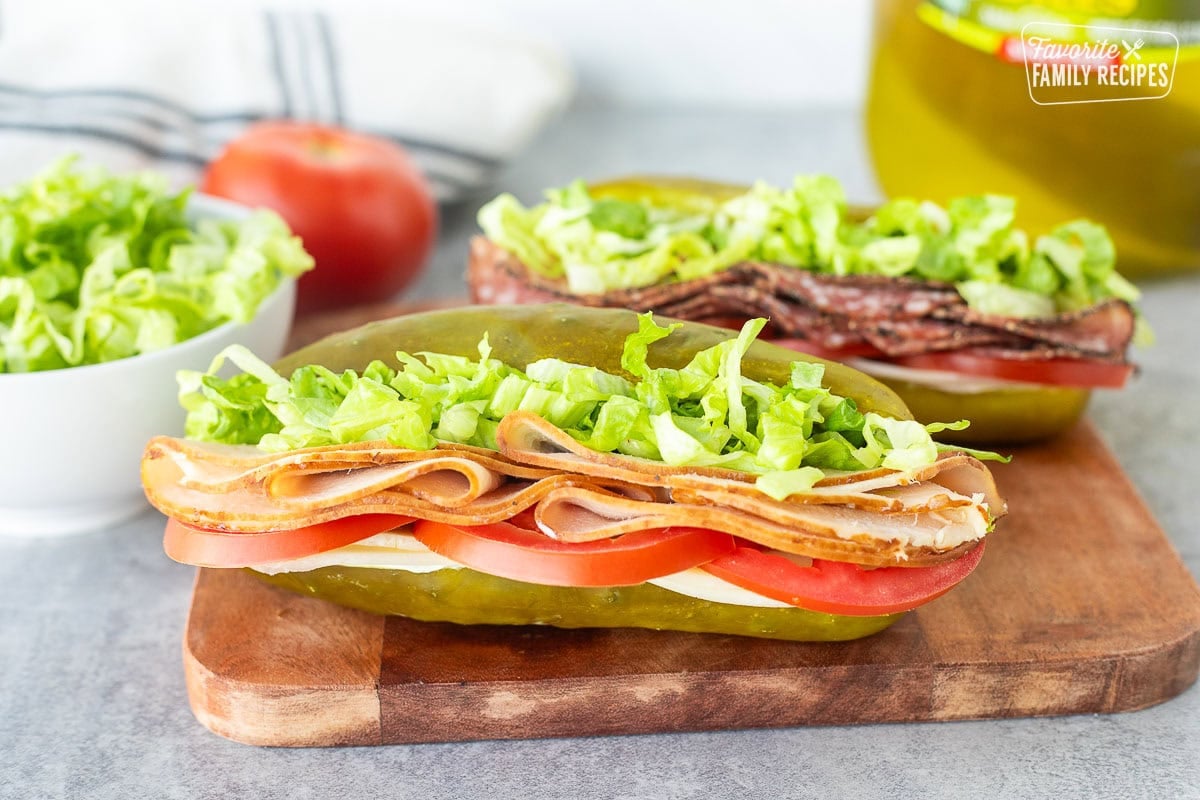 Two pickle sandwiches on a cutting board with turkey, lettuce, tomato, provolone cheese and cream cheese.