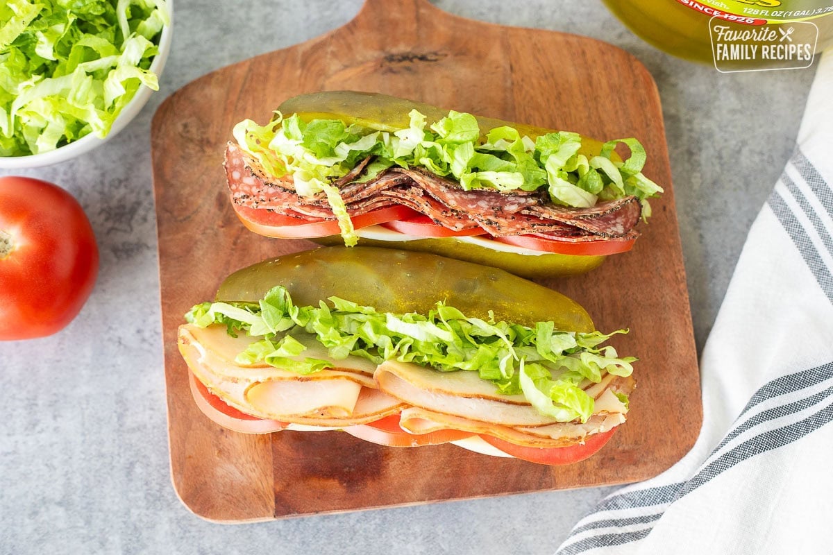 Two pickle sandwiches on a cutting board. One filled with turkey, lettuce, tomato, provolone cheese and cream cheese and the other filled with salami, provolone, lettuce and tomato.