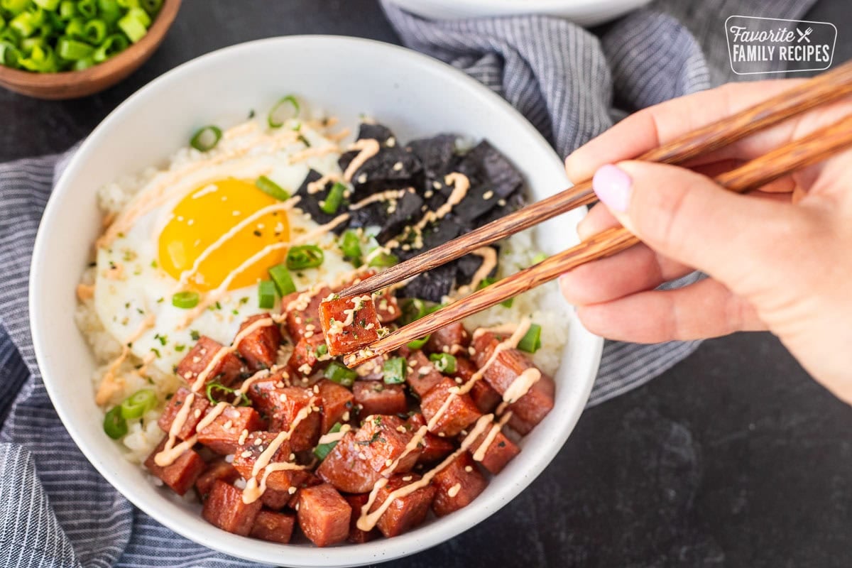 Picking up a piece of spam with chop sticks from a Spam Musubi Bowls with spam, egg, green onion, nori and rice.