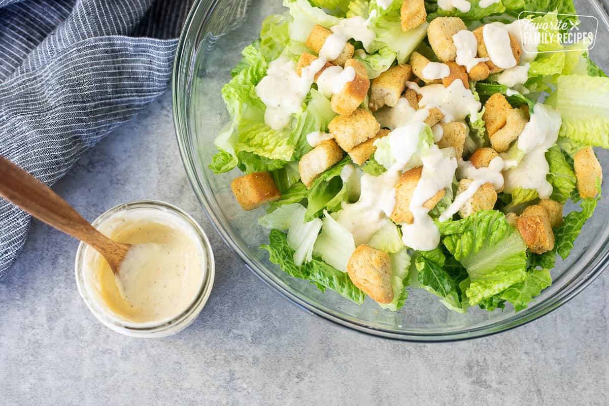 Glass bowl of cut romaine lettuce, croutons and caesar salad dressing on top. Jar of Caesar Salad dressing and spoon on the side.