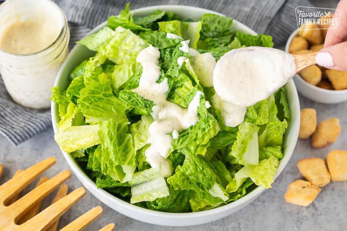 Pouring a spoonful of Caesar Salad Dressing into a bowl of chopped romaine lettuce. Croutons on the side.