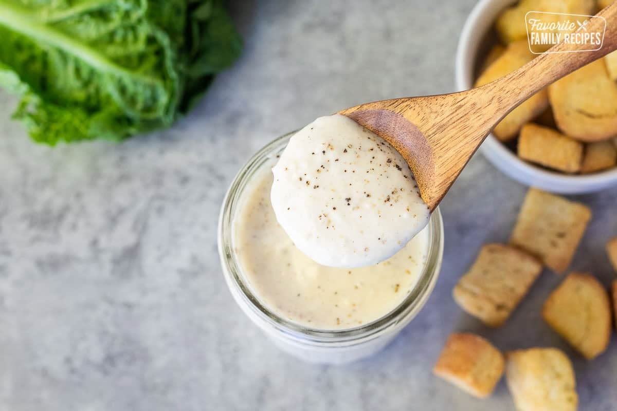 Caesar Salad dressing in a spoon over a jar. romaine lettuce and croutons on the side.