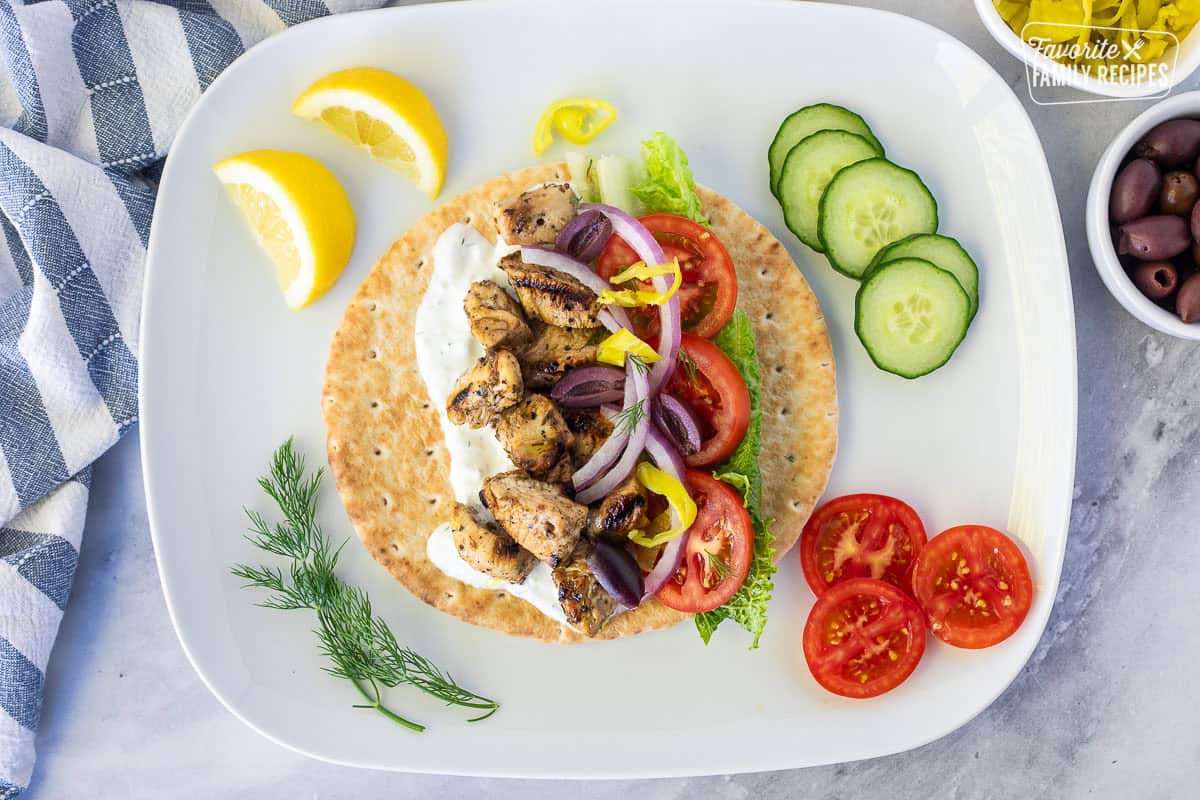 Constructing a Chicken Gyro with pita bread, tzatziki sauce, lettuce, tomato, red onion, Kalamata olives, chicken pieces, pepper and dill.