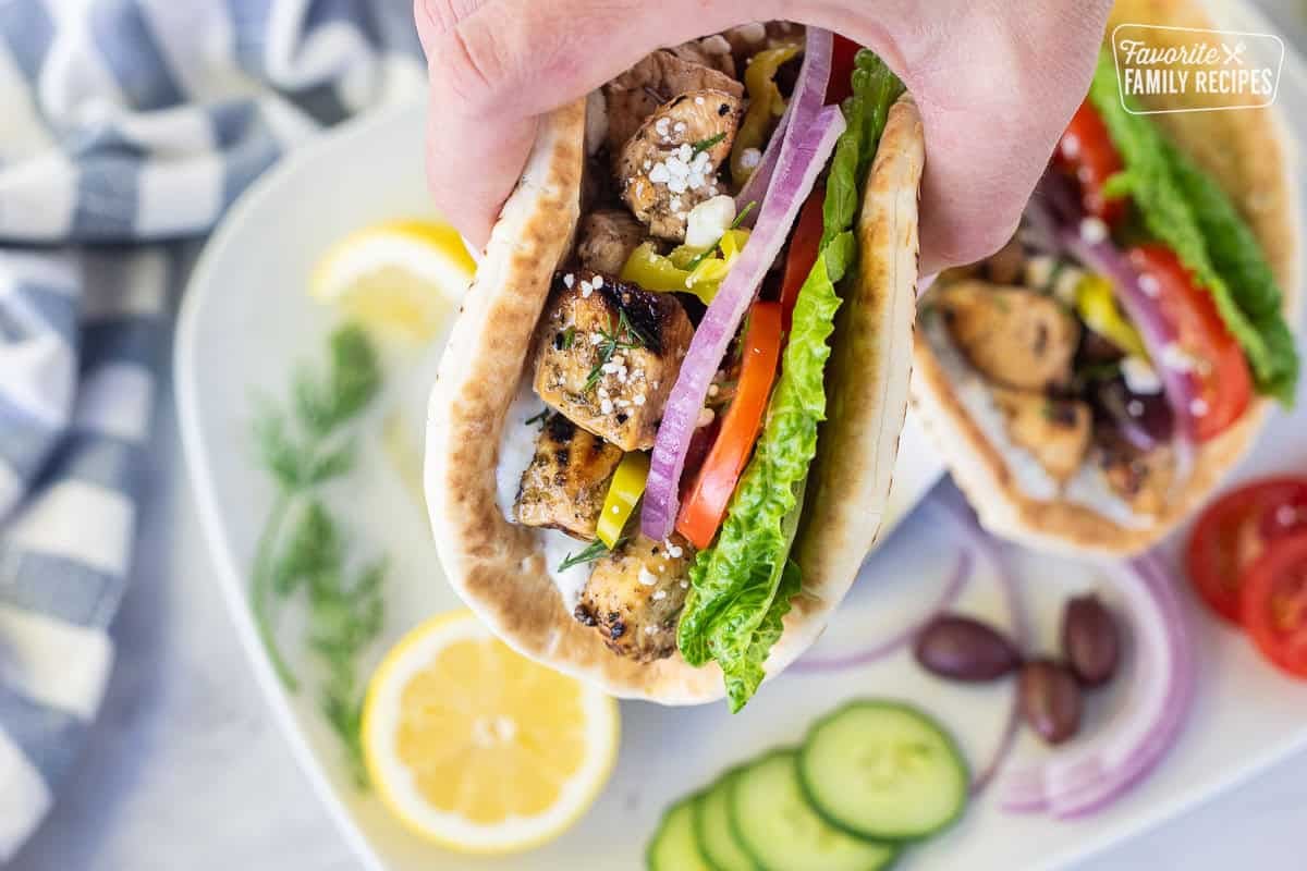 Hand holding a Chicken Gyro with lettuce, tzatziki sauce, chicken, tomatoes, red onions, Kalamata olives, peppers, dill, feta and lemon wedges on the side.