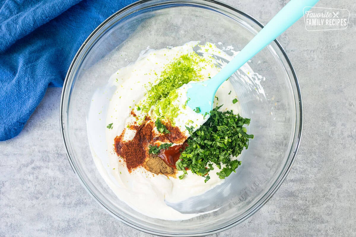 Glass mixing bowl with pasta salad dressing ingredients including mayo, sour cream, cilantro, lime zest and juice, sriracha, chili powder and cumin.