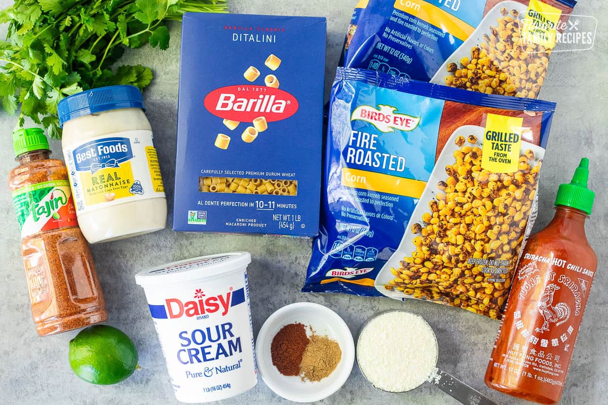 Ingredients to make Elote Pasta Salad including ditalini pasta, corn, sriracha sauce, cojita cheese, tan, chili powder, cumin, sour cream, lime, and mayonnaise.
