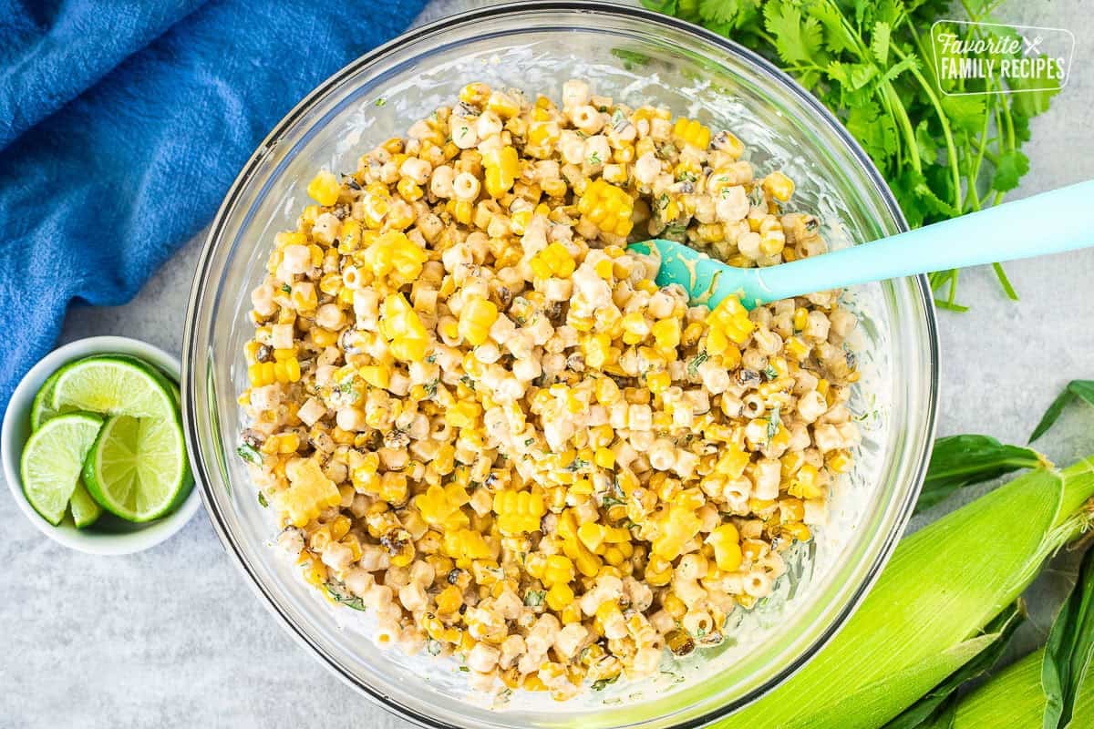 Combining Elote pasta salad in a glass mixing bowl with a spatula.