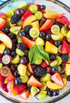 Glass mixing bowl with fresh Fruit Salad including strawberries, pineapple, blackberries, grapes, blueberries, nectarines and kiwis with a simple citrus syrup. Wooden spoon on the side.