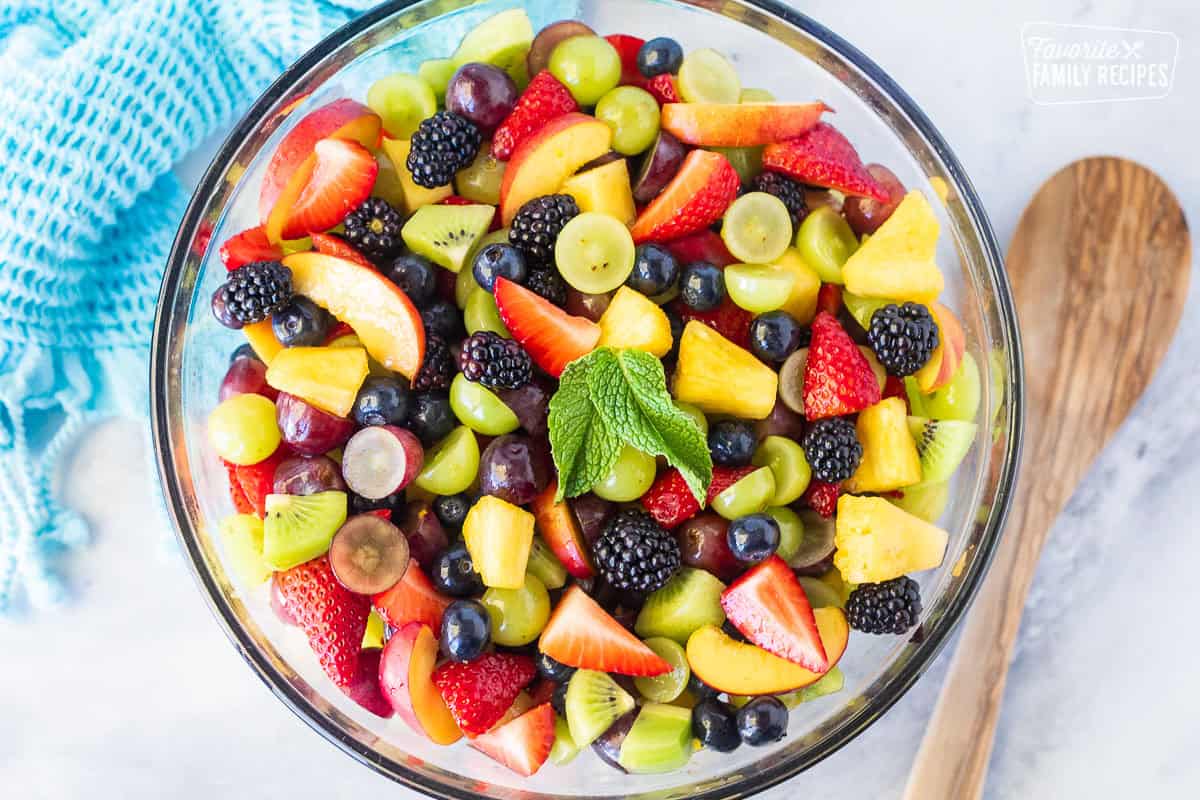 Glass mixing bowl with fresh Fruit Salad including strawberries, pineapple, blackberries, grapes, blueberries, nectarines and kiwis with a simple citrus syrup. Wooden spoon on the side.