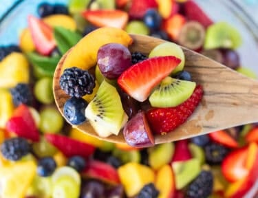Holding up a scoop of Fruit Salad including strawberries, pineapple, blackberries, grapes, blueberries, nectarines and kiwis with a simple citrus syrup.