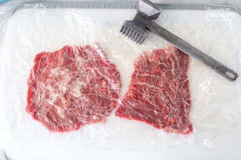 Cutting board with two pieces of skirt steak covered in plastic wrap and flattened with meat tenderizer mallet.