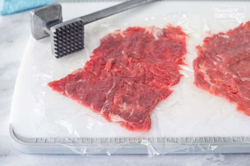 Cutting board with thin pieces of skirt steak on plastic wrap. Mallet resting on the cutting board.